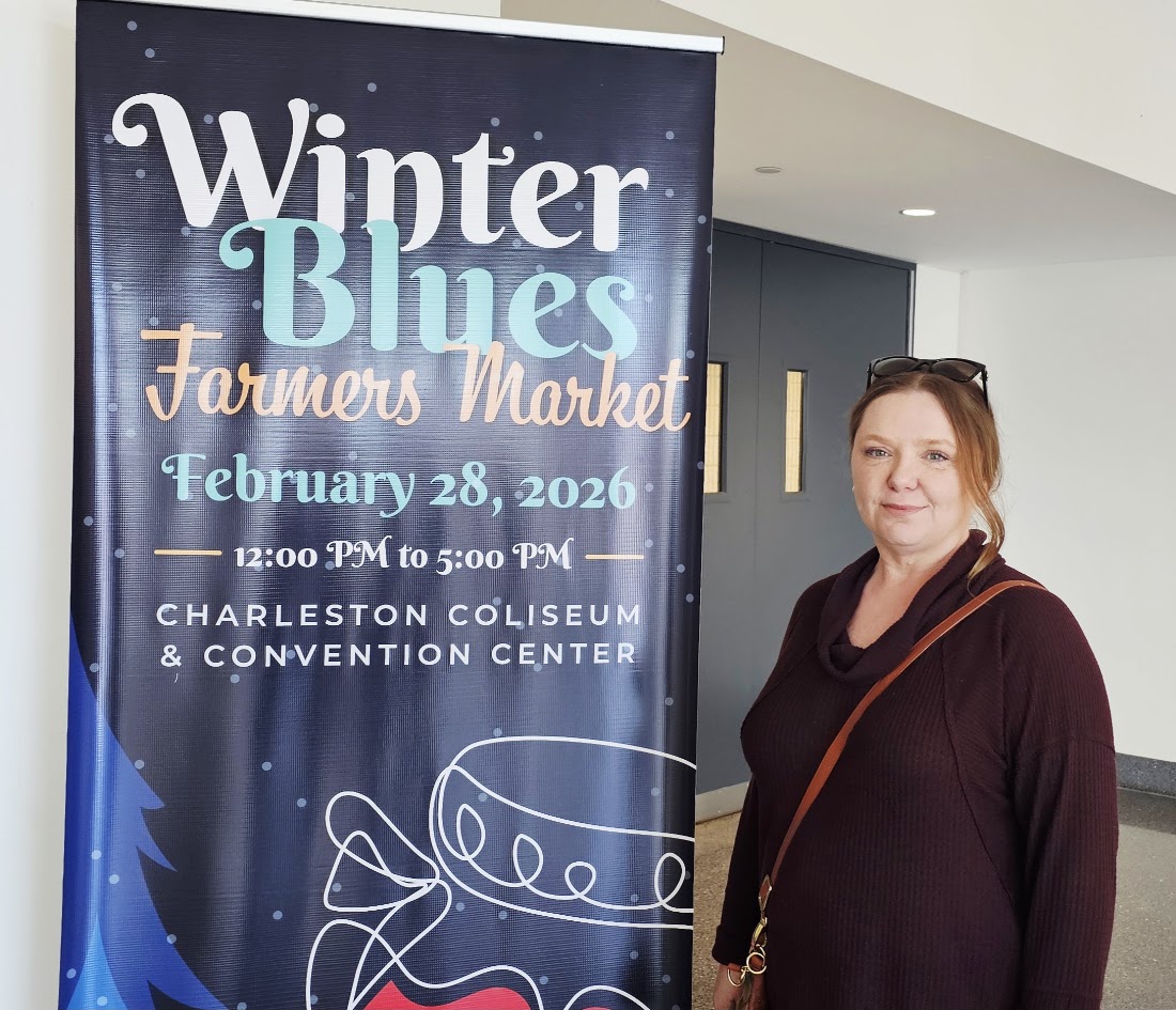 Amy beside the Winter Blues Farmers Market sign at the Charleston Coliseum and Convention Center.